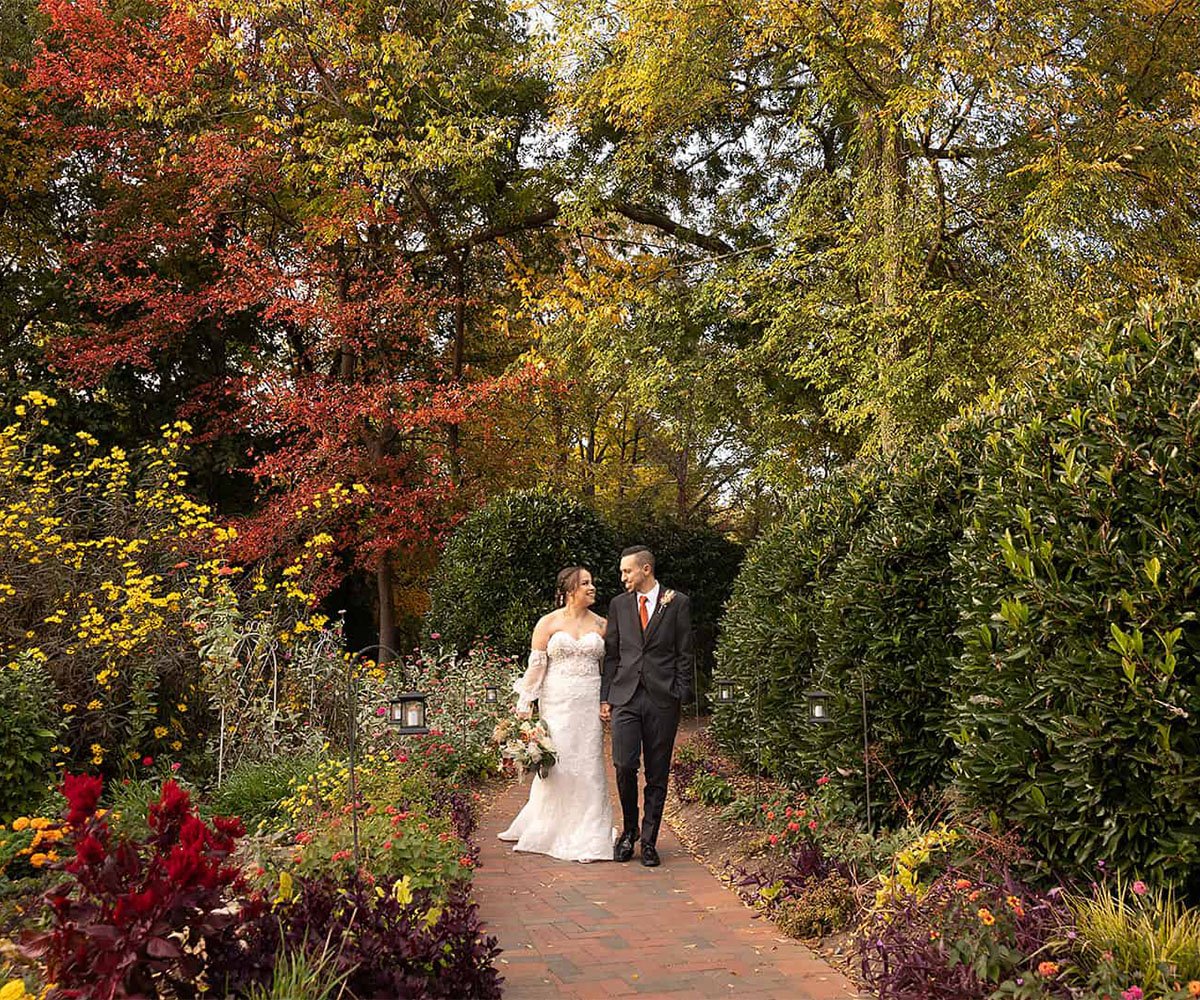 Newlyweds strolling through vibrant autumn garden pathway at Alexander Homestead wedding venue in Charlotte NC with colorful fal