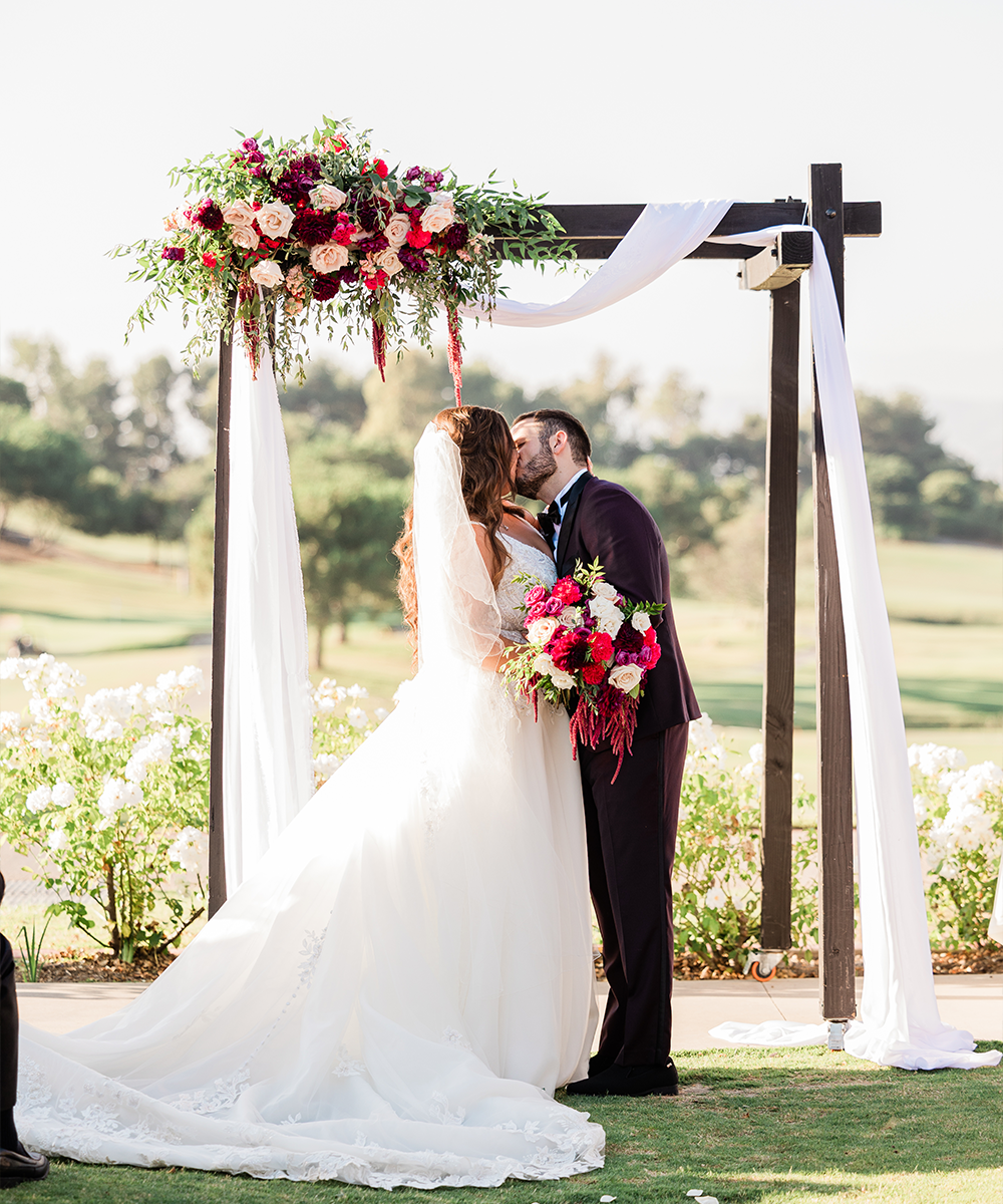 Couple kissing at arbor lawn arch with red flowers - Aliso Viejo by Wedgewood Weddings