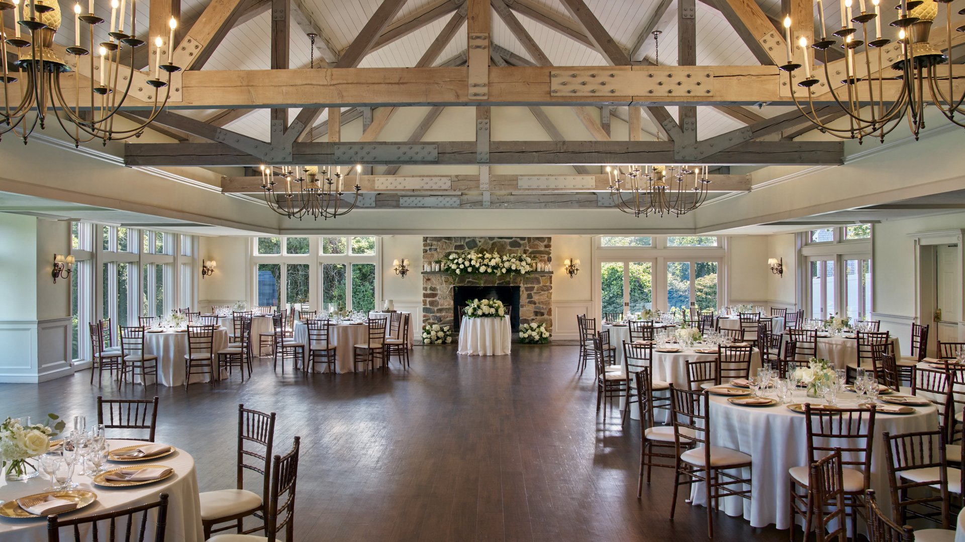 The Williams Room at The Barker House beautifully set for a wedding reception with round tables, white linens, gold chargers, and floral centerpieces under rustic wood beams and chandeliers.