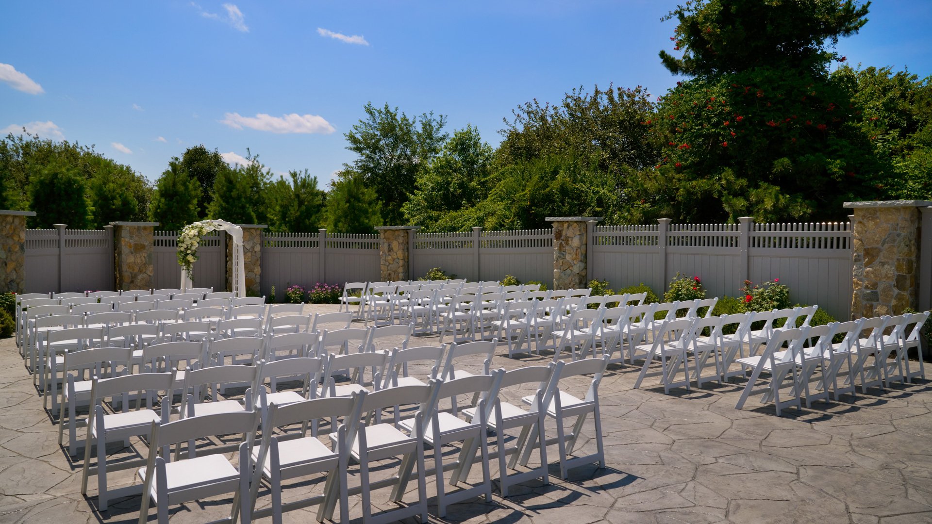 Charming outdoor wedding ceremony at The Barker House garden, with white chairs, stone fencing, and a floral arch beneath a bright blue sky.