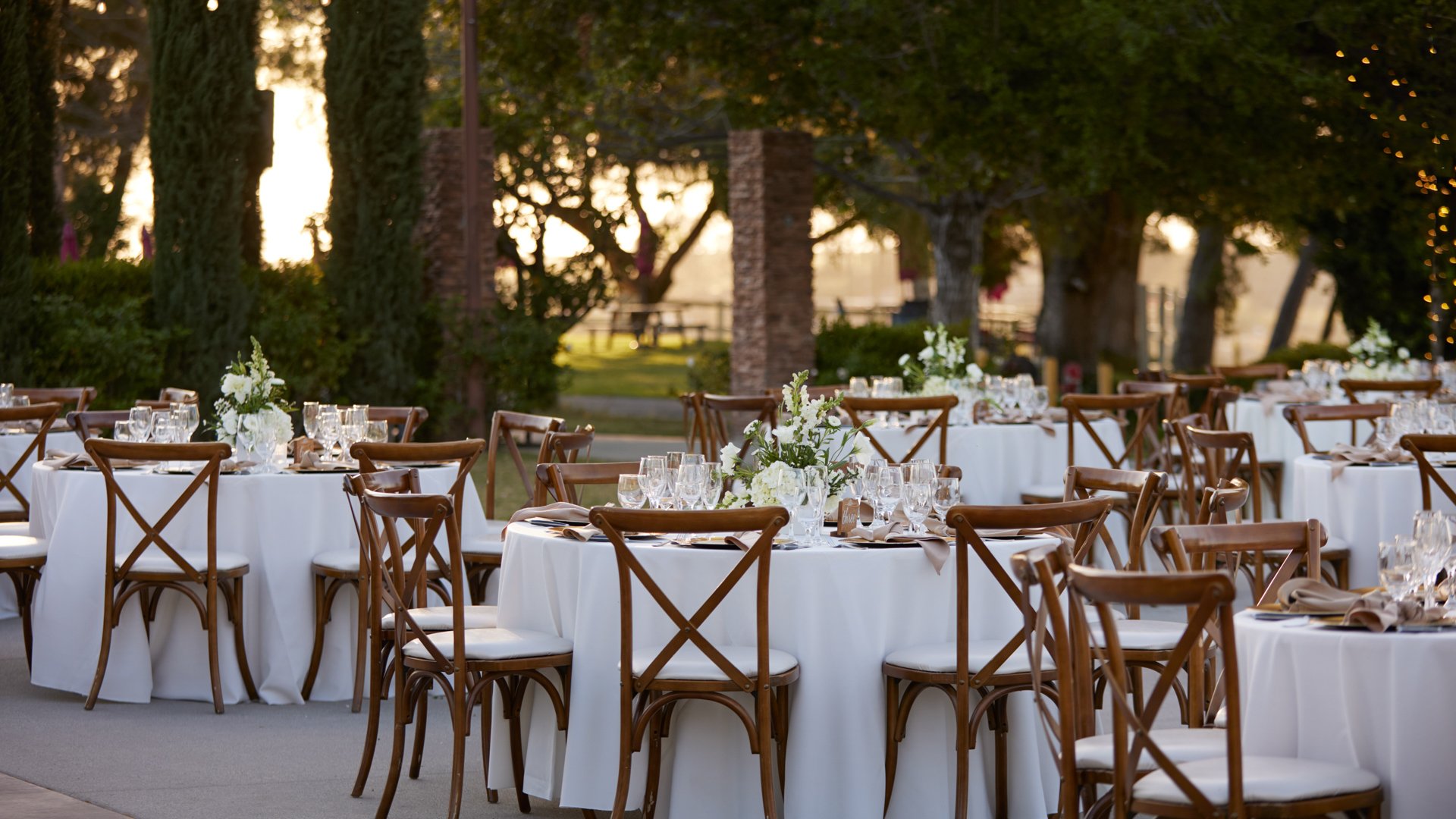 Round reception tables with white linens and cross-back chairs at Bel Vino Winery courtyard during golden hour.