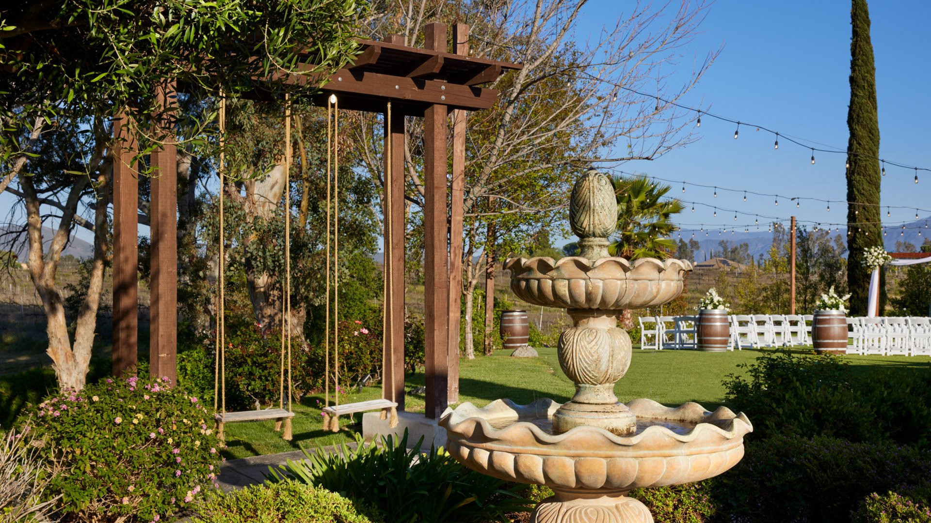 Stone fountain and wooden swing set on the cocktail patio at Bel Vino Winery with vineyard views and ceremony lawn in the background.