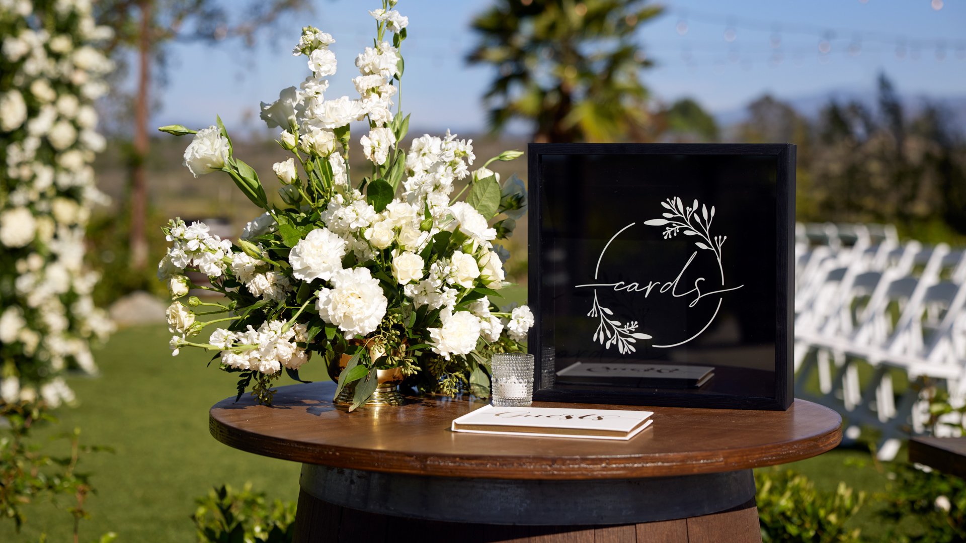 Elegant welcome table setup at Bel Vino Winery featuring floral arrangement, guest card box, and white ceremony seating in the background.