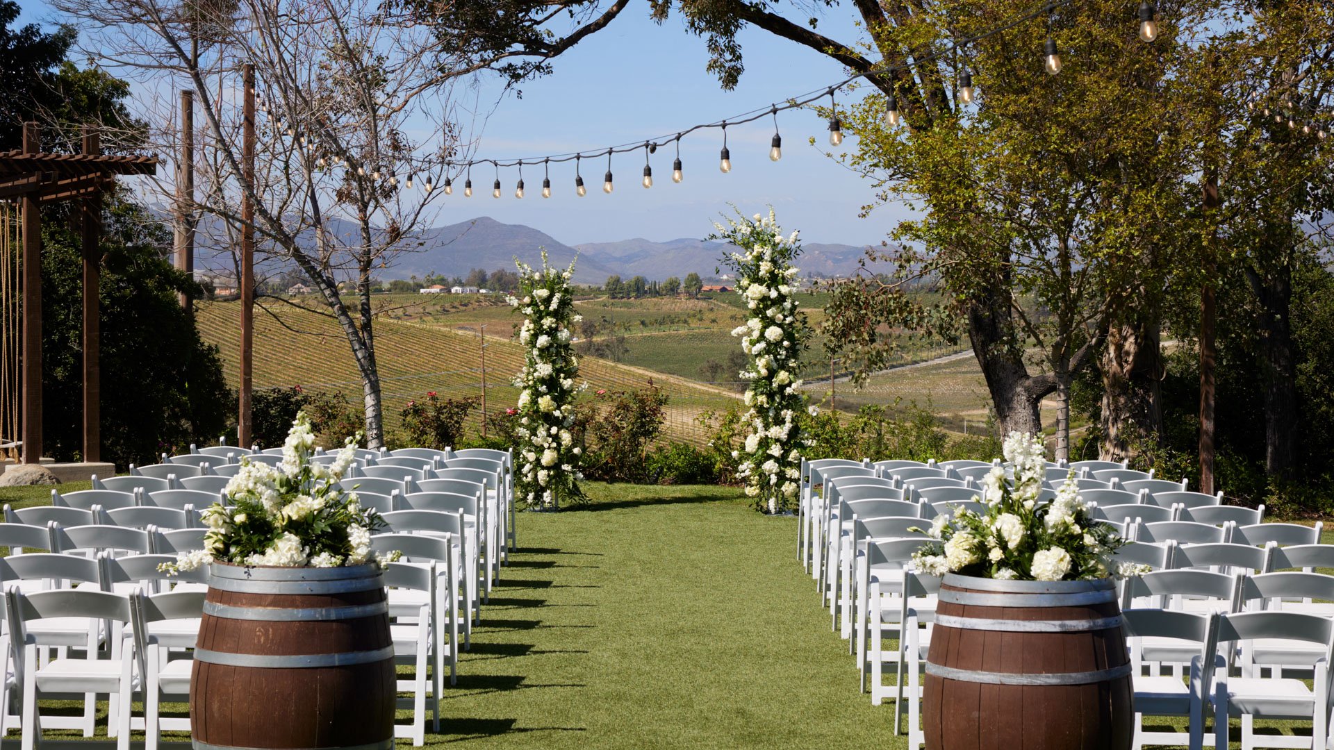 Ceremony aisle at Bel Vino Winery lined with white chairs and floral wine barrels overlooking Temecula vineyards.