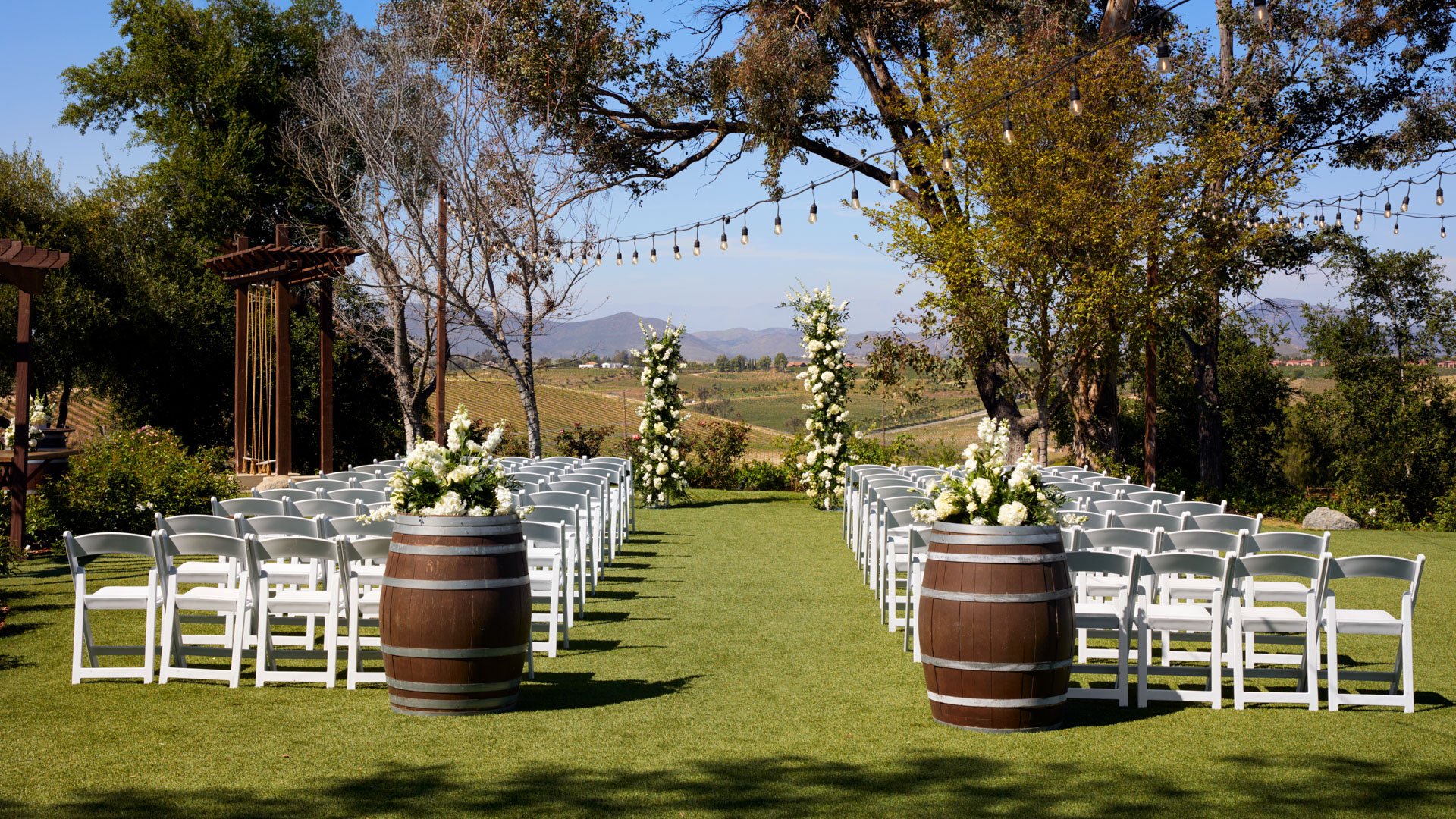 Rustic ceremony setup at Bel Vino Winery with white chairs, floral barrels, and sweeping vineyard landscape.