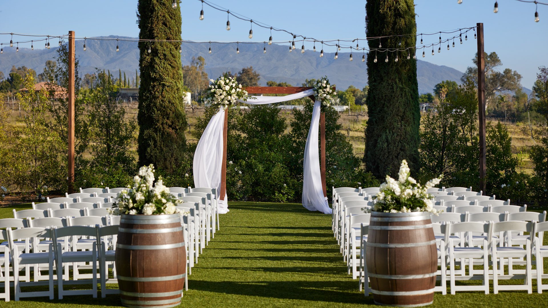 Scenic wedding ceremony at Bel Vino Winery featuring wine barrels, white florals, and panoramic vineyard backdrop.