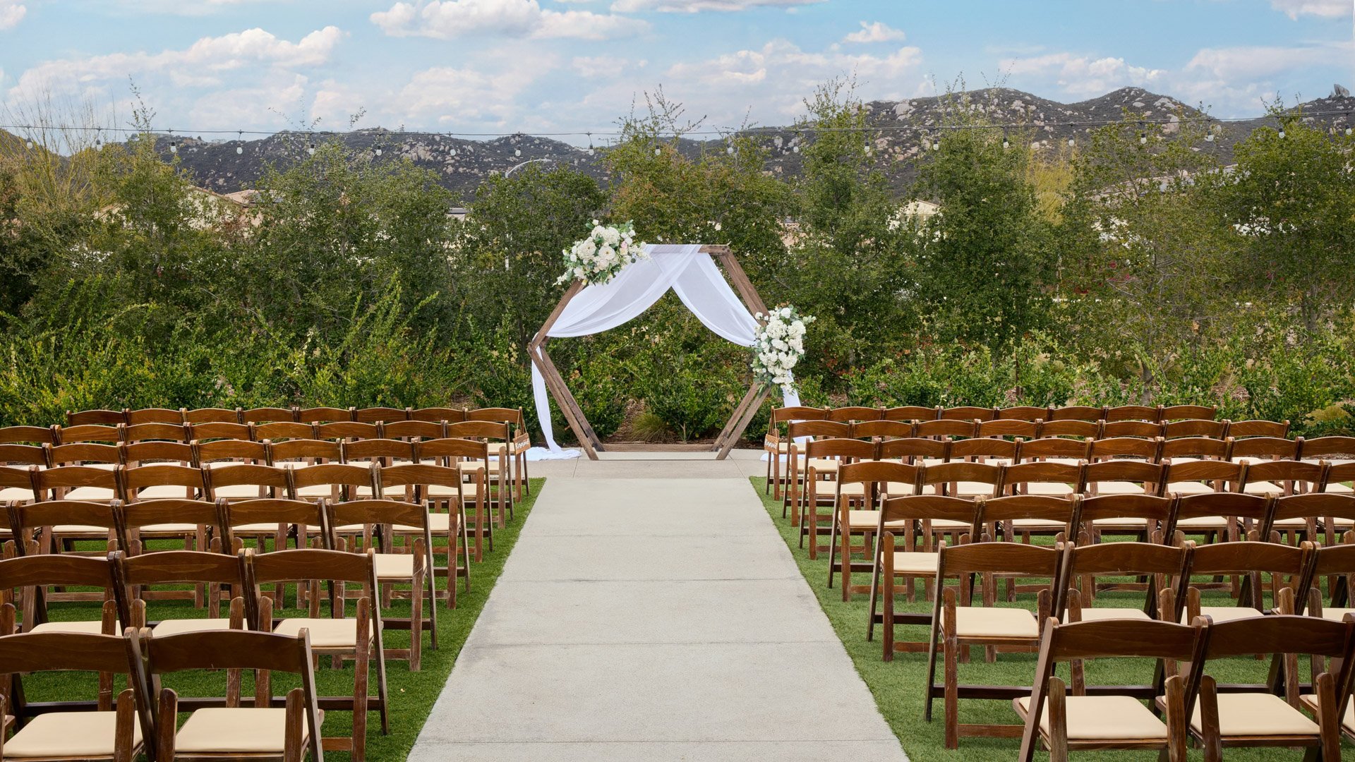 Wooden chairs lined along an outdoor ceremony aisle leading to a hexagon arch draped in white fabric and florals at Canopy Grove wedding venue in Escondido, California.
