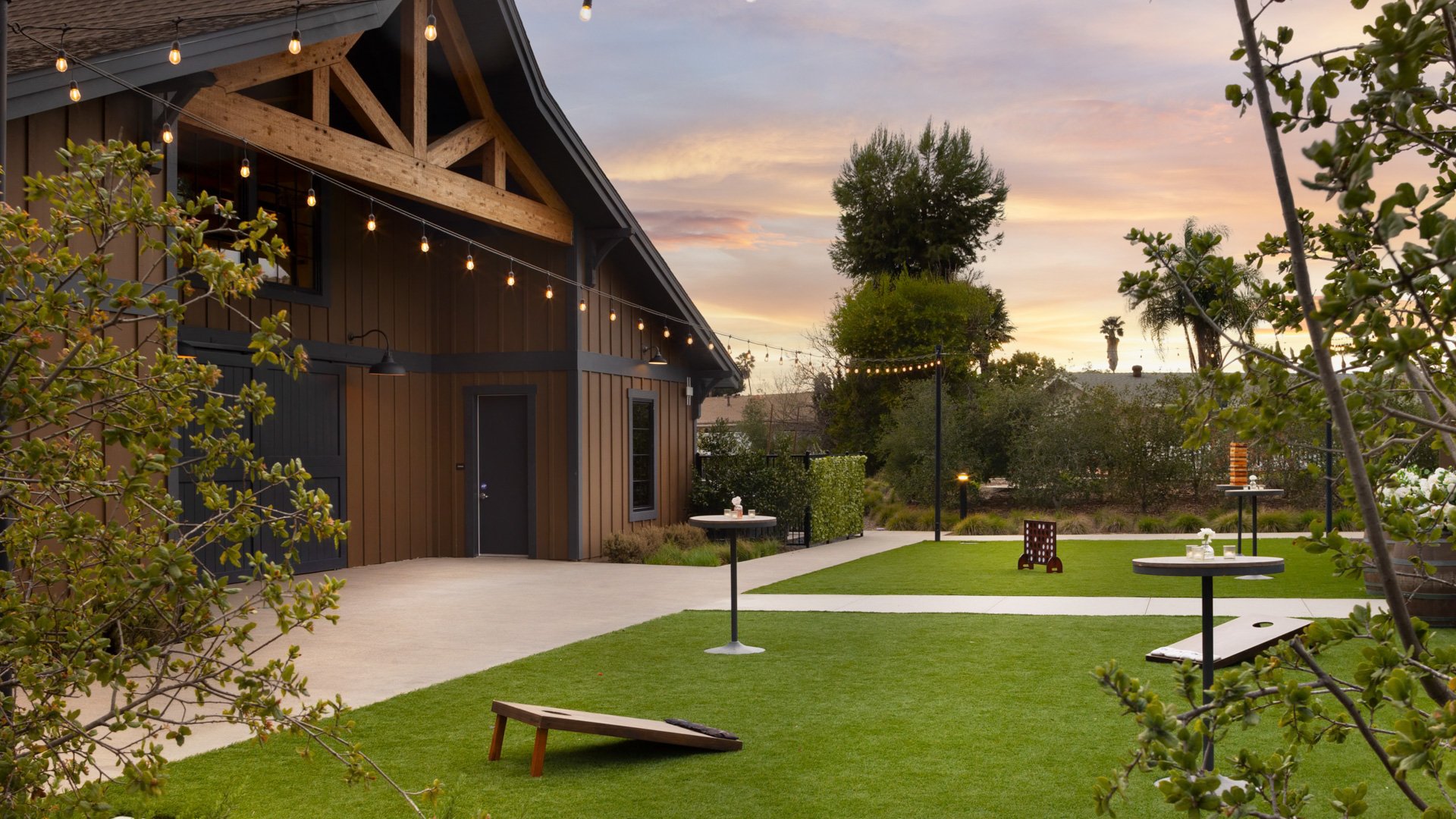 Evening view of Canopy Grove’s outdoor cocktail patio with cornhole, string lights, and cocktail tables beside the rustic barn