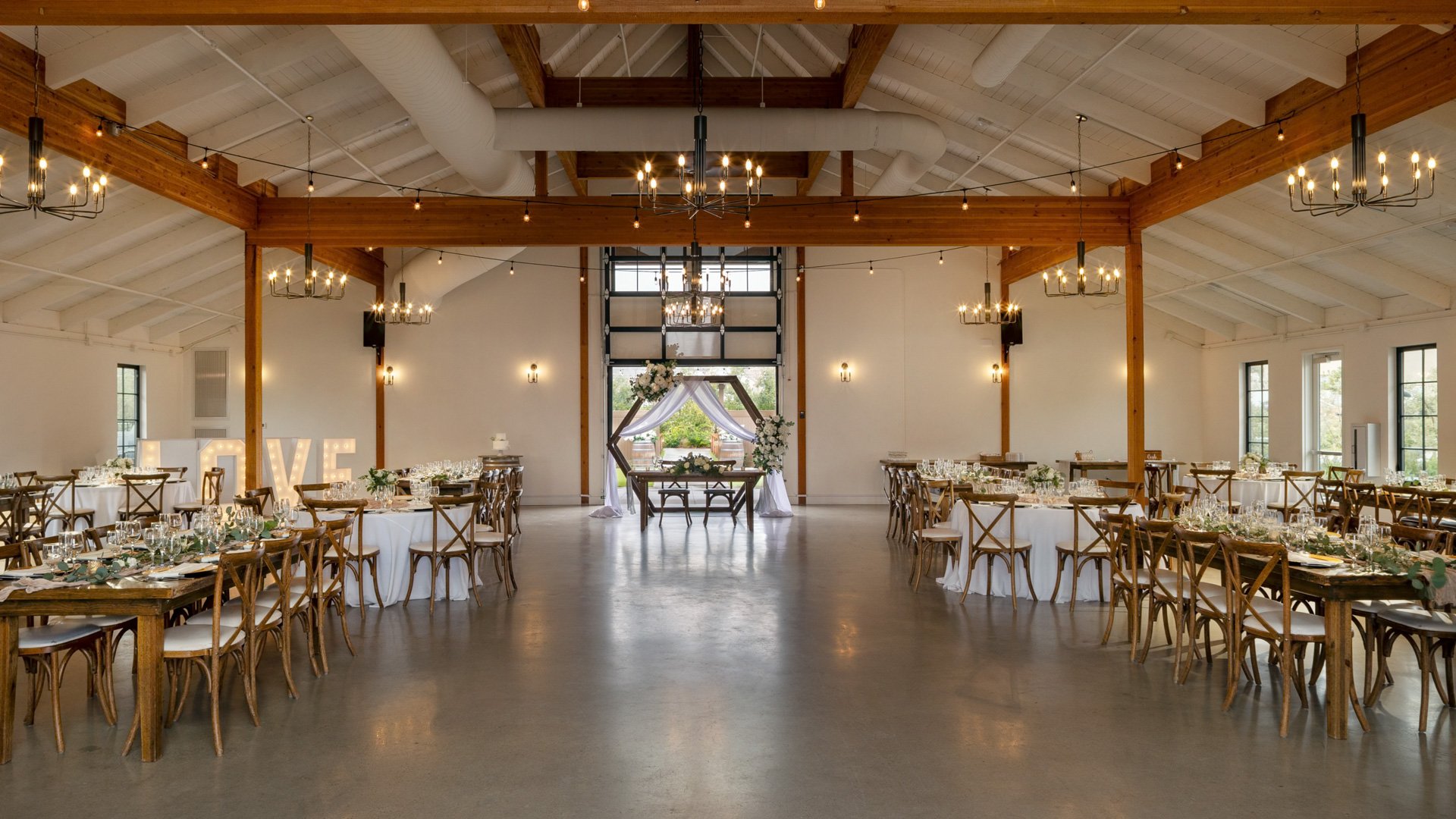 Spacious wedding barn at Canopy Grove featuring rustic chandeliers, wood beams, and a view of the outdoor ceremony arch through the open doors.