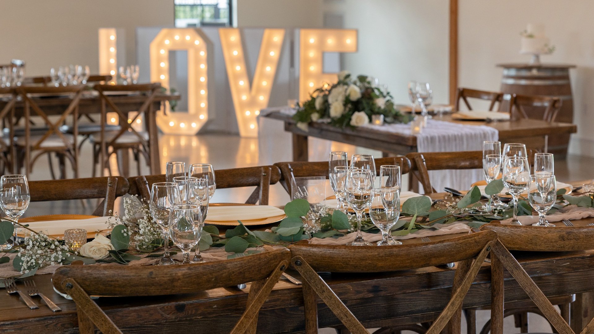 Close-up of rustic wood tables with greenery runners and glassware, with a large illuminated LOVE sign in the background at Canopy Grove wedding venue.