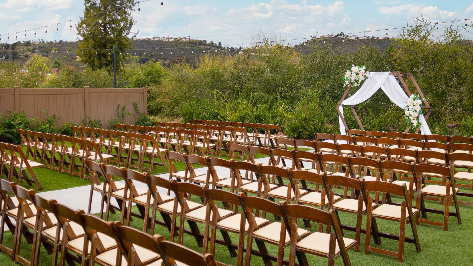 Side view of rows of wooden chairs facing a decorated ceremony arch on the outdoor lawn at Canopy Grove in Escondido, CA.