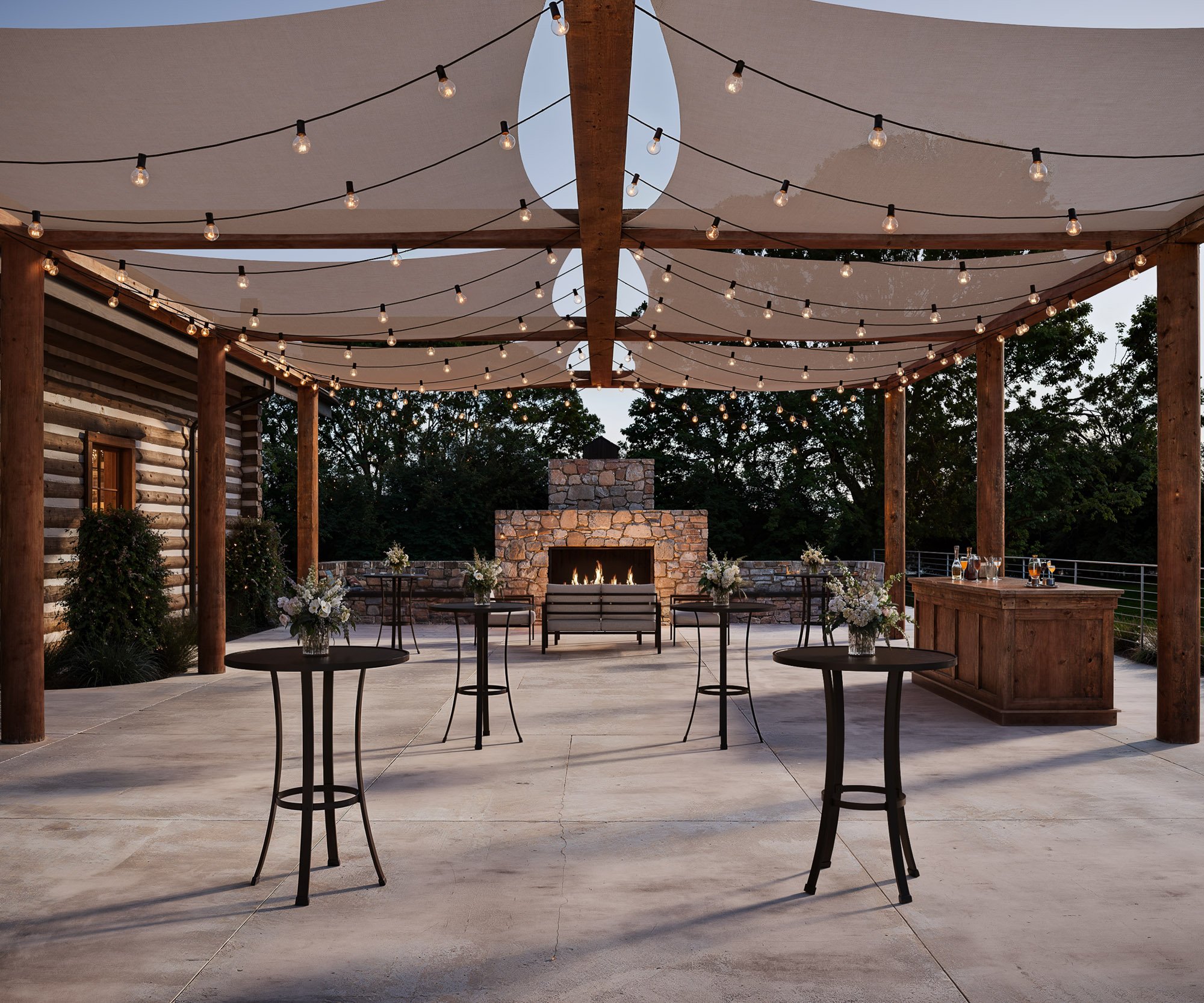 Charming outdoor cocktail area featuring wooden pergola with string lights, stone fireplace, high-top tables with floral arrangements, and wooden bar at Casa del Rio.