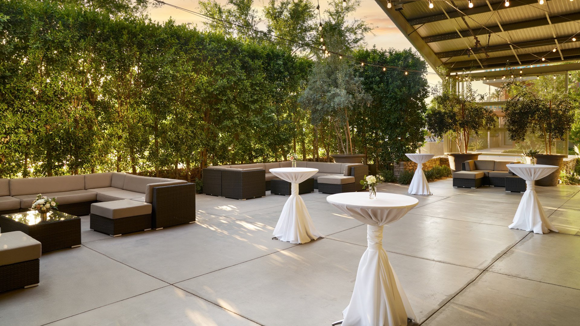 Outdoor cocktail lounge at The Clayton House in Scottsdale, AZ, featuring modern wicker seating, cocktail tables draped in white linens, and romantic string lights surrounded by lush greenery.