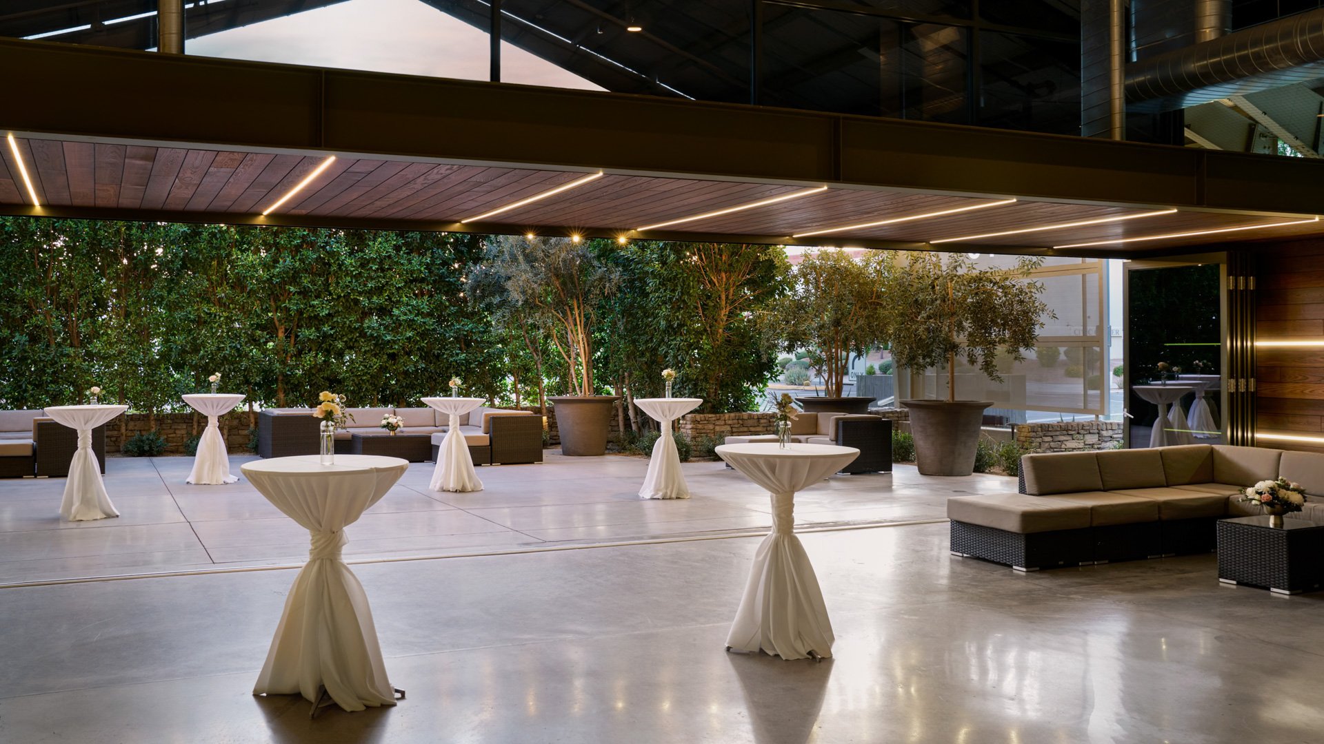 Atrium space at The Clayton House in Scottsdale, AZ, set for cocktail hour with high-top tables, white drapery, and cozy lounge seating beneath a covered patio with greenery views.
