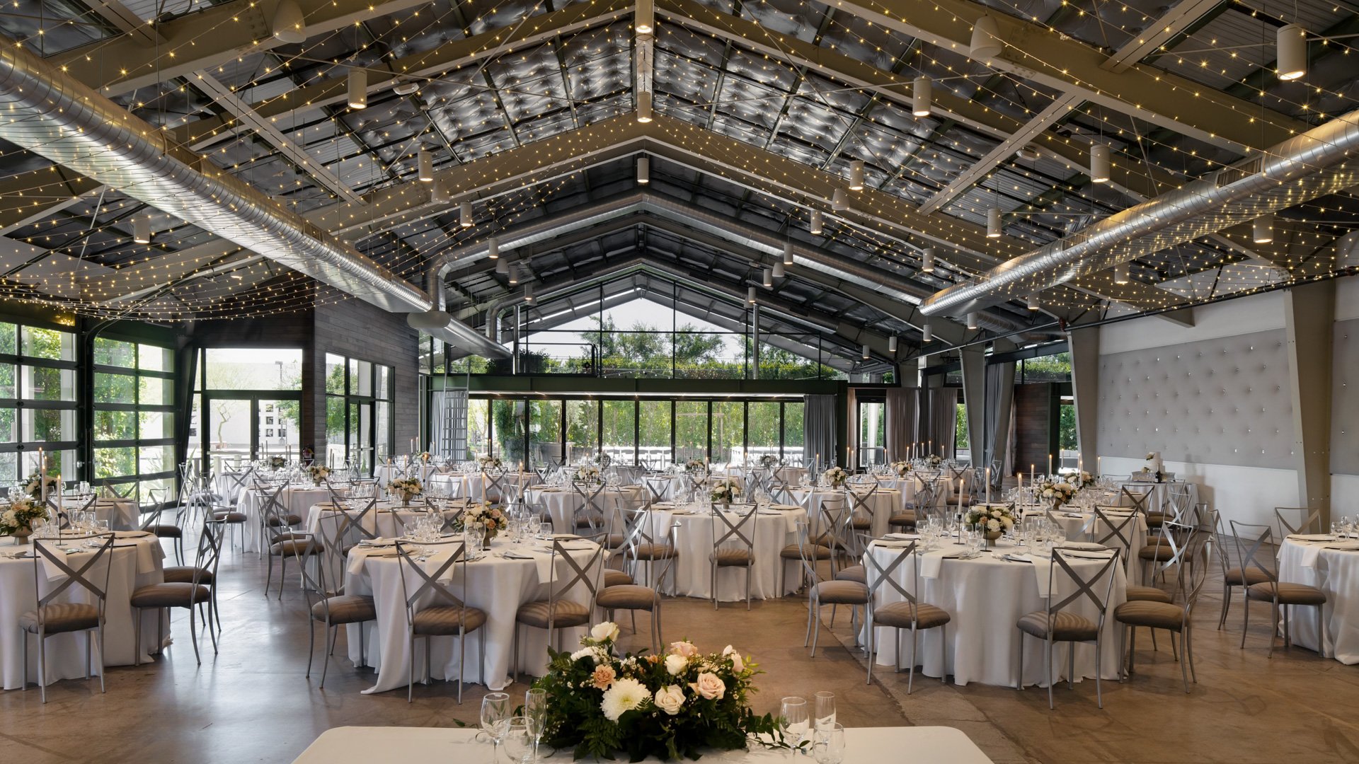 Reception setup inside The Hangar at The Clayton House, featuring white round tables, neutral décor, string lighting, and open glass walls creating an airy and elegant wedding ambiance.