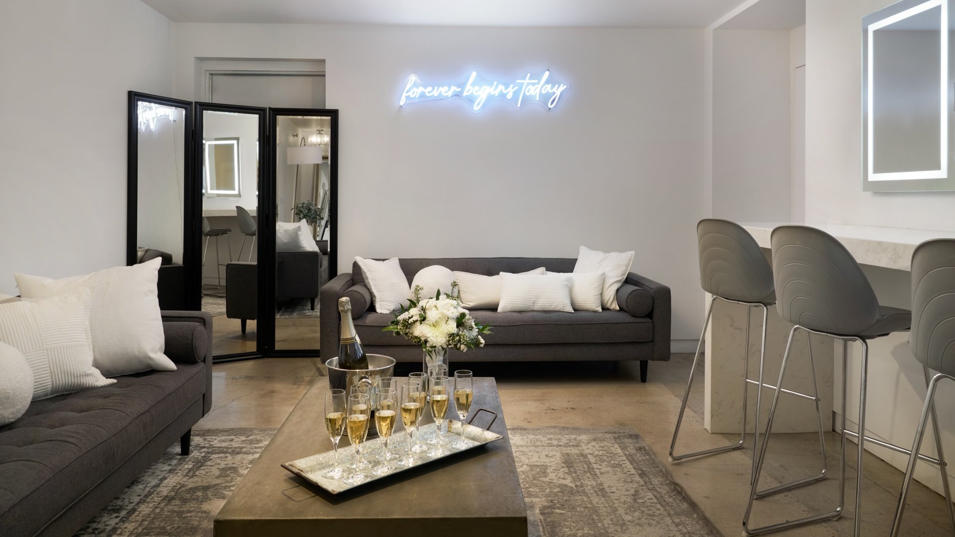Wedding suite at The Clayton House in Scottsdale, AZ, featuring plush gray sofas, illuminated vanity mirrors, and a neon “Forever Begins Today” sign — an elegant space for couples to relax and get ready.