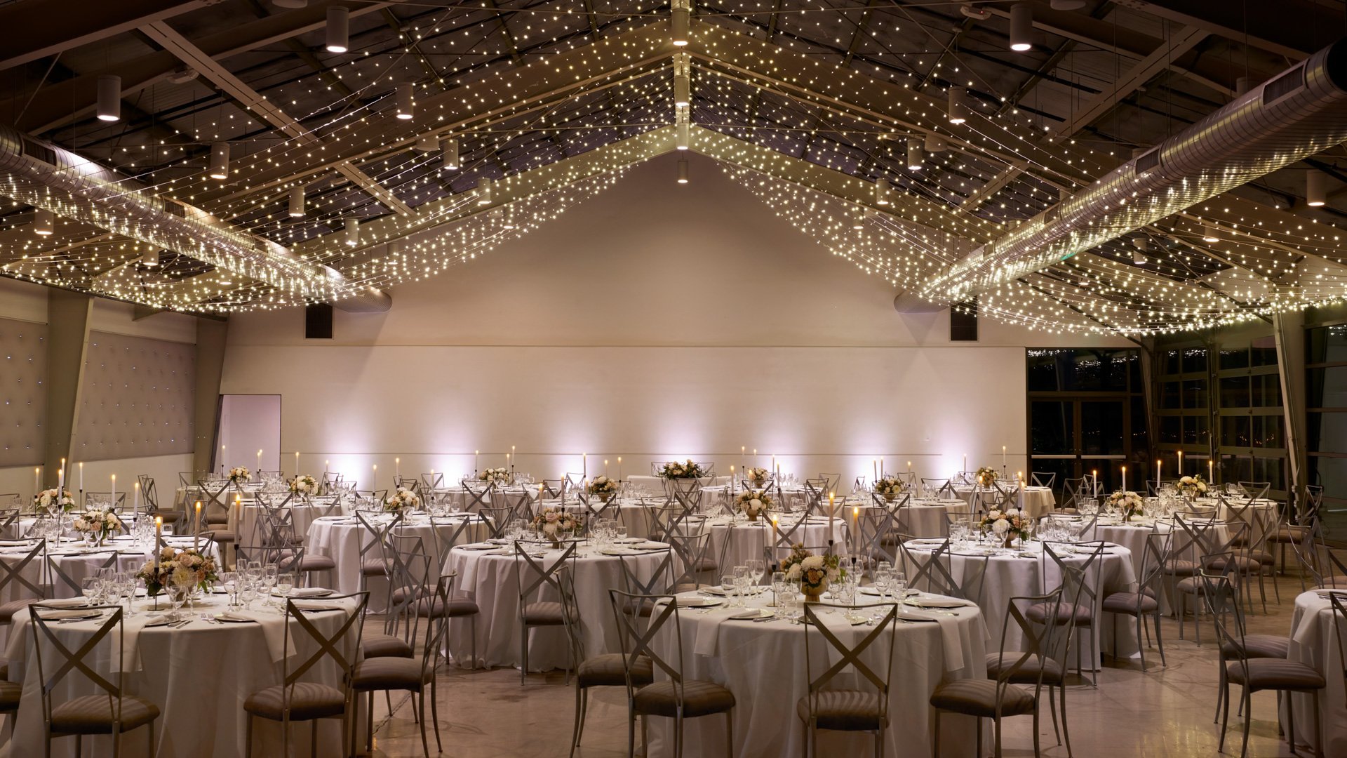 Reception setup inside The Hangar at The Clayton House in Scottsdale, AZ, showcasing round tables with white linens, candle centerpieces, and twinkle-light ceiling décor for a glamorous modern wedding.