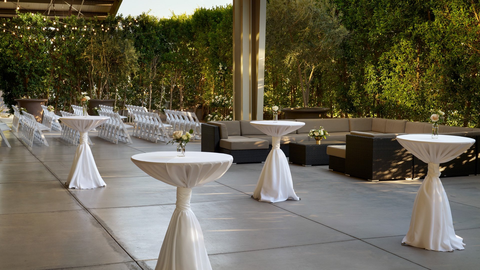 Outdoor cocktail and ceremony patio at The Clayton House in Scottsdale, Arizona, with white cocktail tables, lounge seating, floral accents, and romantic string lights surrounded by greenery.