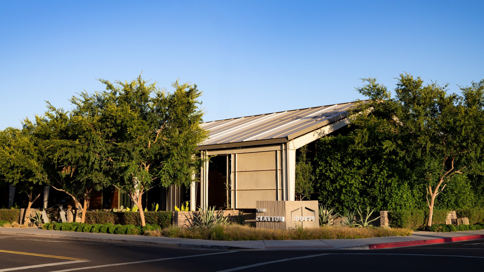 Exterior daytime view of The Clayton House in Scottsdale, Arizona, showing modern industrial architecture framed by lush trees and desert landscaping under a clear blue sky.