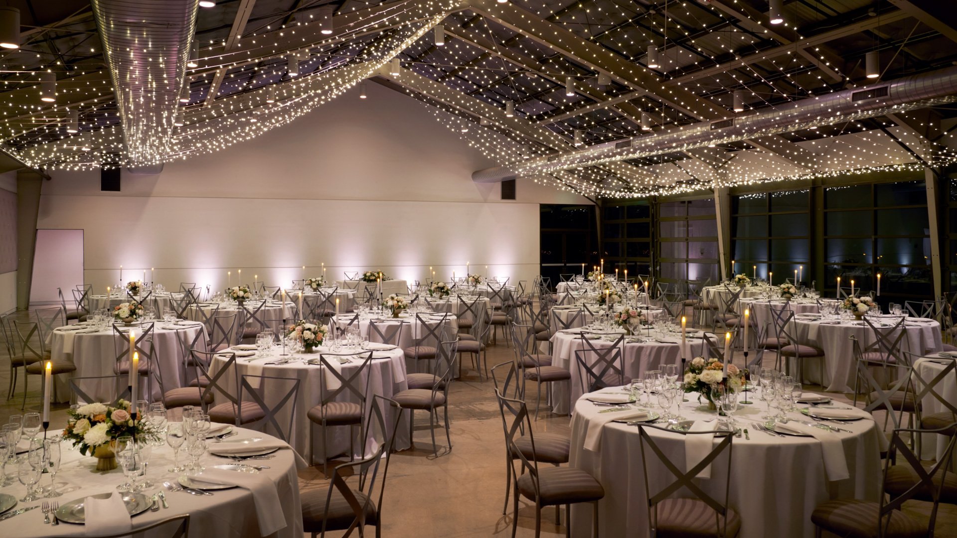 The Hangar reception hall at The Clayton House in Scottsdale, AZ, elegantly decorated with white round tables, floral centerpieces, glowing candles, and twinkle lights draped across the ceiling.