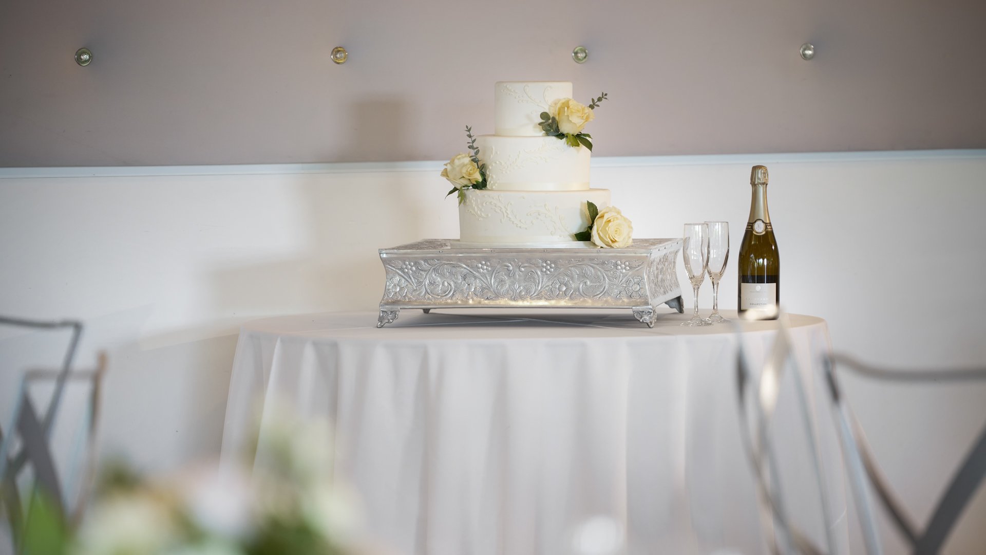 Close-up of a tiered white wedding cake with ivory roses on a silver stand, displayed at The Clayton House in Scottsdale, Arizona, with champagne and glasses set for the celebration.