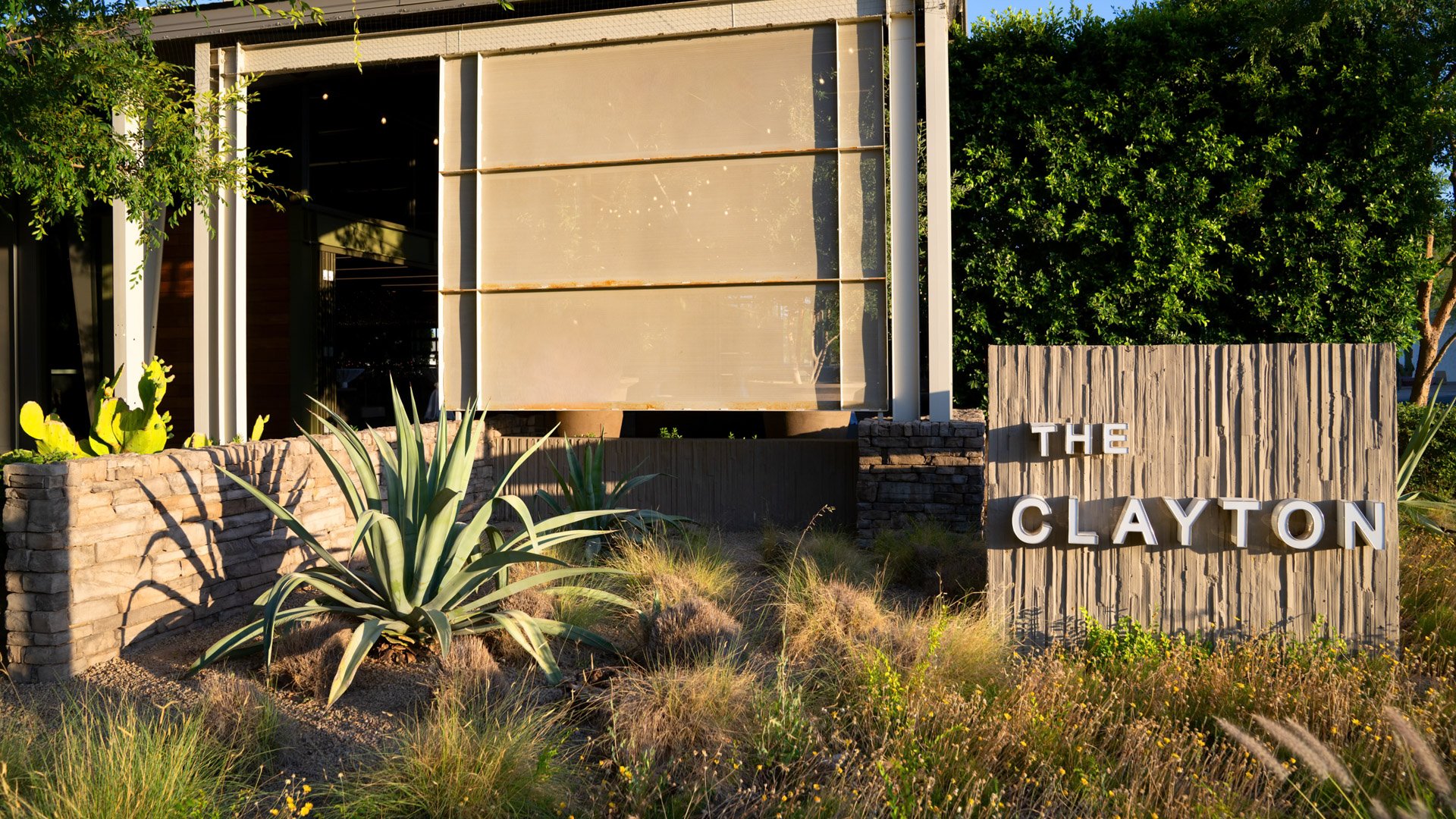Front signage of The Clayton House in Scottsdale, Arizona, featuring modern lettering, stone accents, and desert plants like agave and cacti illuminated by the afternoon sun.