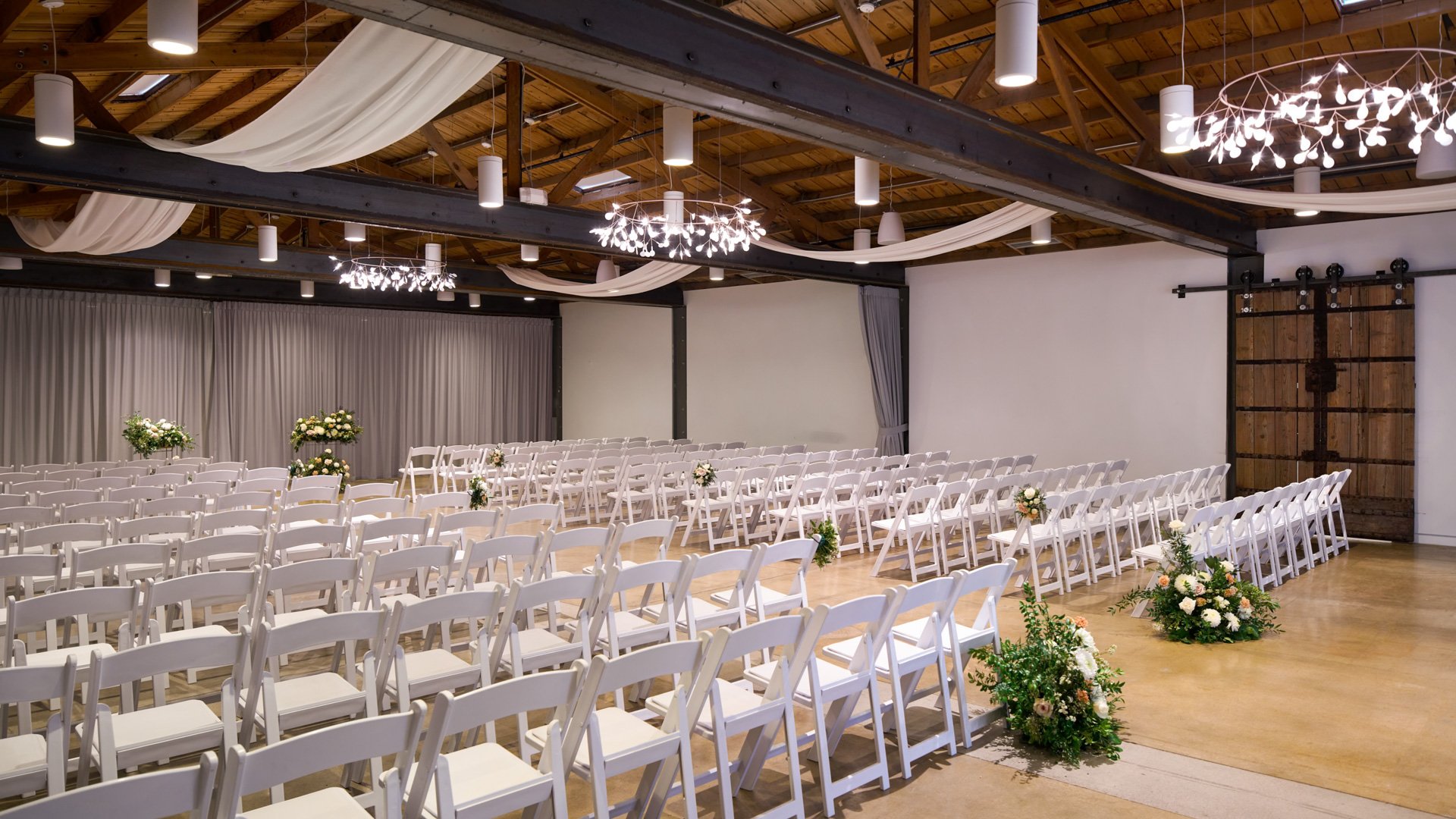 Indoor ceremony space called The Rafters at The Clayton House in Scottsdale, Arizona, with white chairs, soft draping, floral décor, and warm lighting beneath exposed wooden beams—ideal for a modern yet cozy wedding.