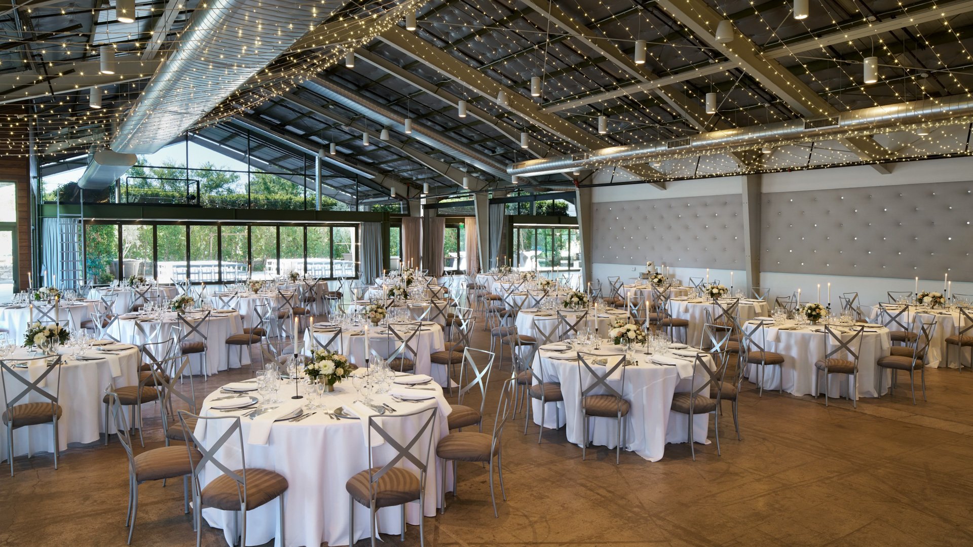 The Hangar ballroom at The Clayton House in Scottsdale, AZ, prepared for a wedding reception with round tables, soft candlelight, floral centerpieces, and ceiling draped in glowing string lights.