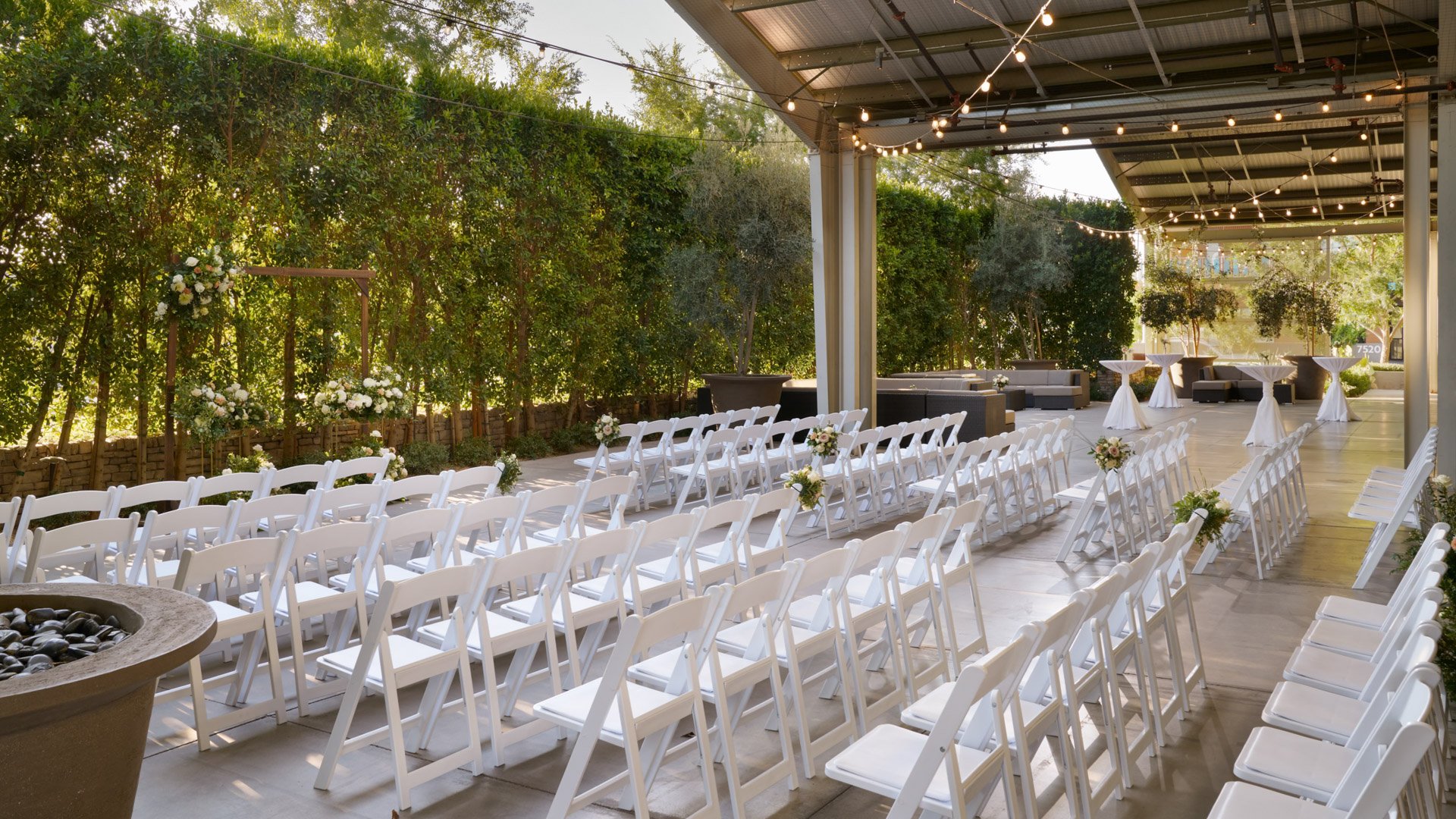 Romantic outdoor ceremony at The Clayton House, featuring white chairs, greenery-lined aisles, and a wooden arch adorned with ivory roses and lush foliage beneath café string lights.
