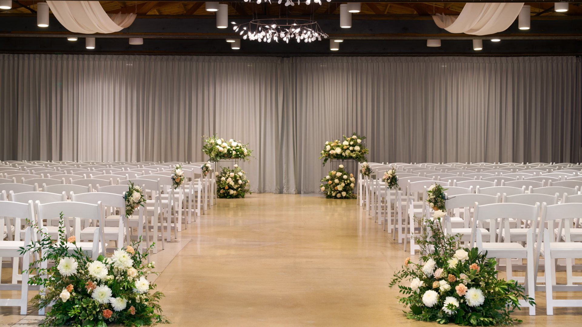 Indoor ceremony at The Clayton House’s Rafters room in Scottsdale, AZ, showcasing rustic wood beams, soft white drapery, and elegant floral arrangements lining the aisle for a cozy wedding setting.