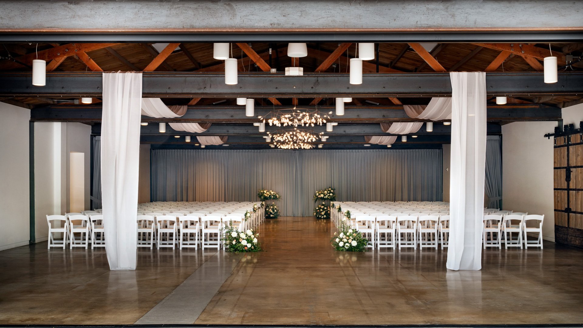 Wide view of The Rafters at The Clayton House in Scottsdale, Arizona, featuring white ceremony chairs, hanging chandeliers, draped ceiling fabric, and floral décor at the altar for a timeless indoor wedding.