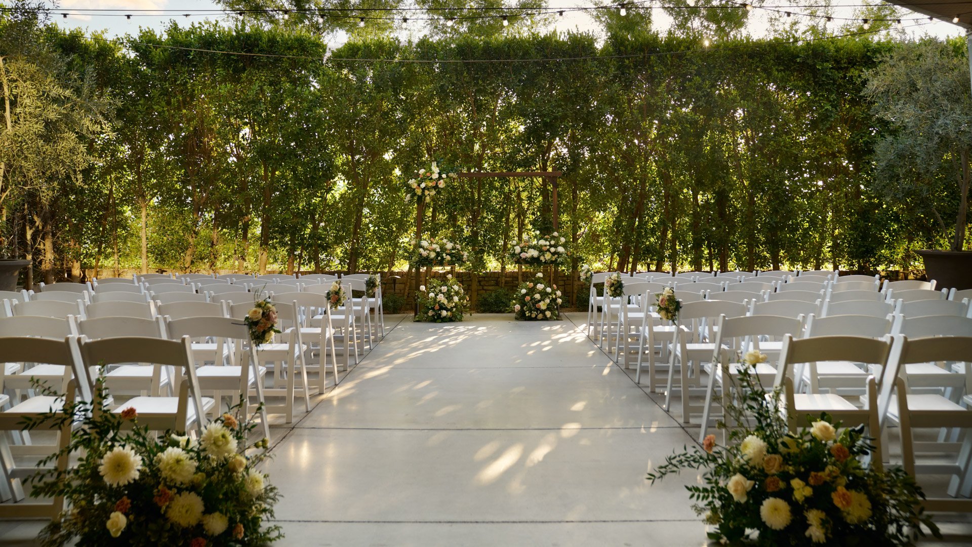 Serene outdoor wedding ceremony setup at The Clayton House Porch in Scottsdale, Arizona, with white seating, romantic floral aisle décor, and a wood arch backdrop framed by sunlit greenery and string lights.