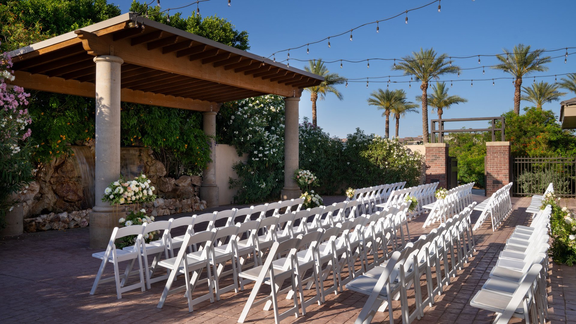 Grand Waterfall courtyard ceremony setup at Colby Falls Arizona with white chairs, stone waterfall backdrop, palm trees, and floral