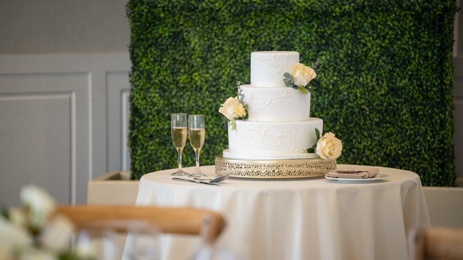 Three-tier white wedding cake with floral accents on a gold stand in the Colby Falls Grand Hall reception space in Arizona.