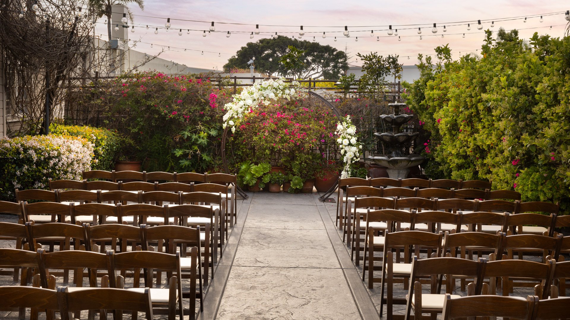 Elegant outdoor wedding ceremony setup in the Garden Courtyard at Cuvier Club by Wedgewood Weddings in La Jolla, featuring wooden chairs arranged for guests with lush bougainvillea blooms and string lights overhead