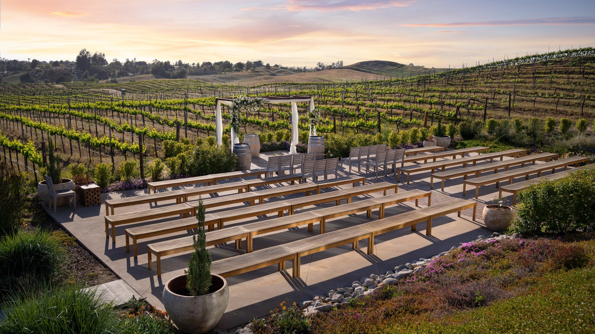 Outdoor vineyard wedding ceremony site at Danza del Sol Winery in Temecula, California, featuring rustic benches, floral arch, and panoramic vineyard views at sunset.