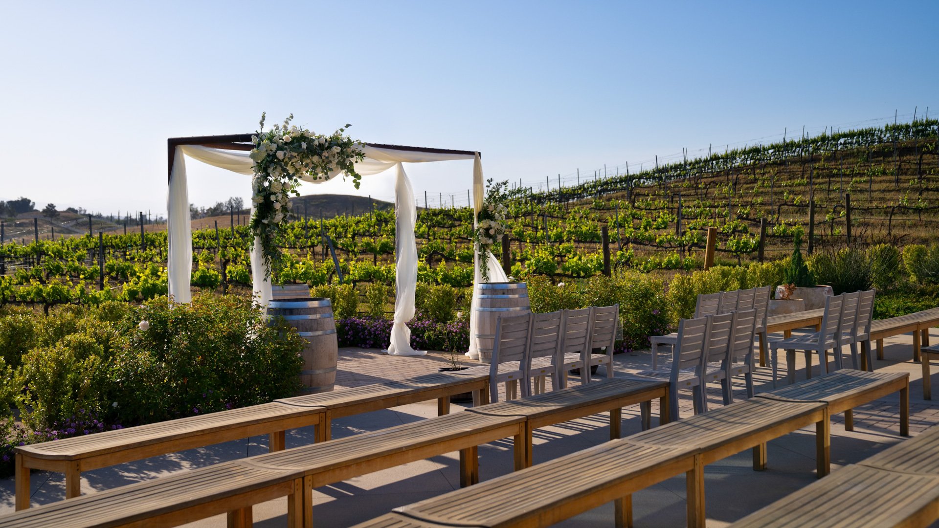 Danza del Sol Winery wedding ceremony site in Temecula, California, featuring a floral arch, wine barrels, and vineyard hillside backdrop.