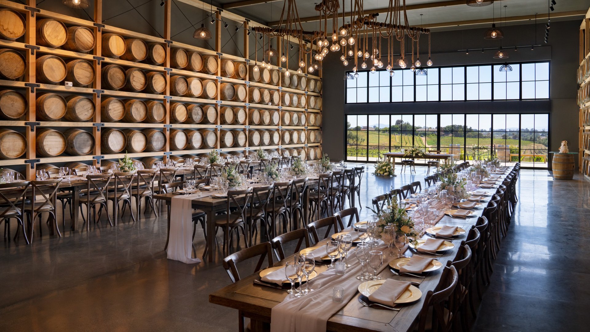Elegant winery wedding reception in the Barrel Room at Danza del Sol Winery in Temecula, CA, with long rustic tables, wine barrel wall, and hanging Edison bulb chandeliers.