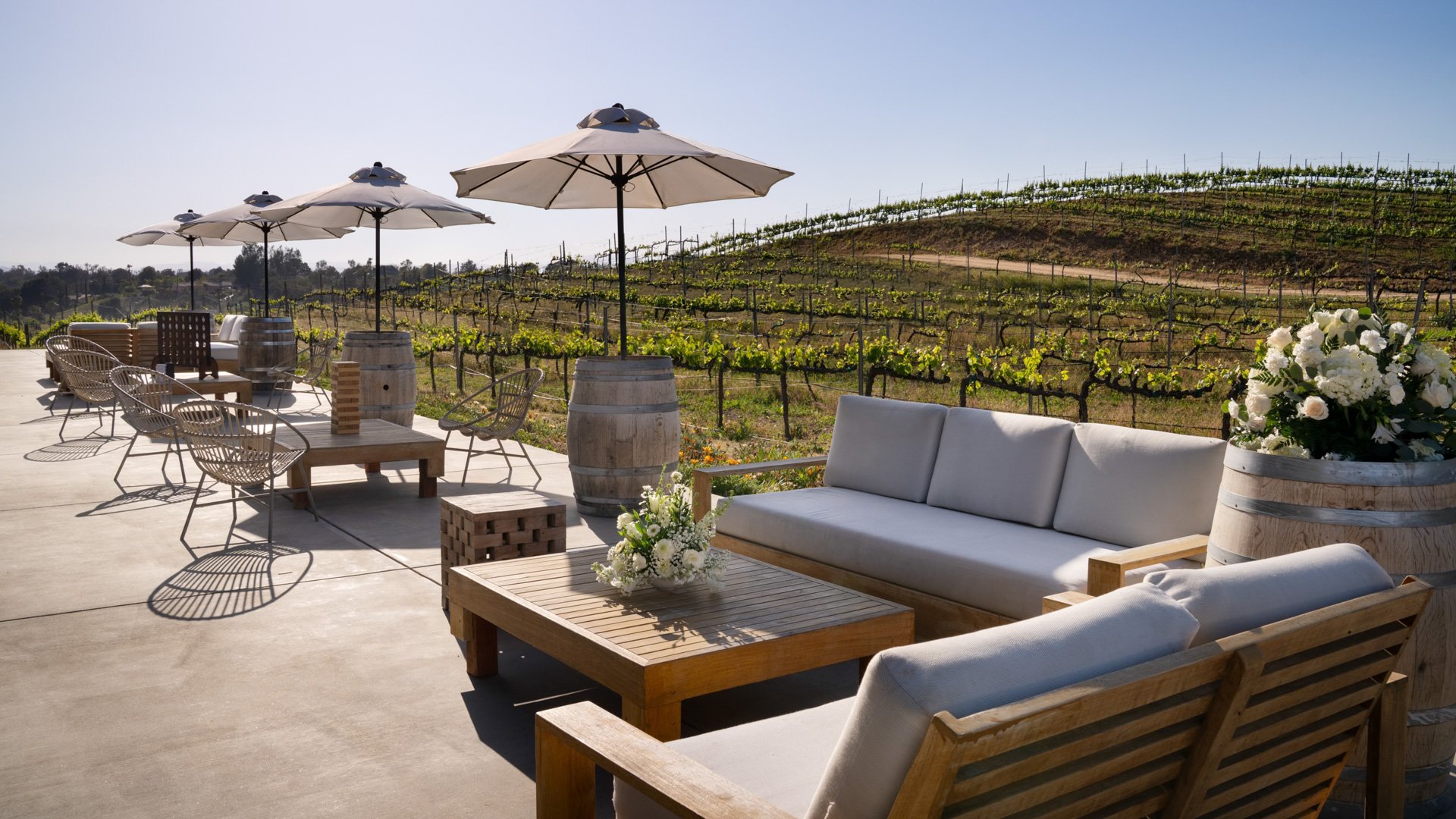 Outdoor cocktail lounge at Danza del Sol Winery in Temecula, CA, with vineyard backdrop, cozy lounge seating, umbrellas, and wine barrel tables.