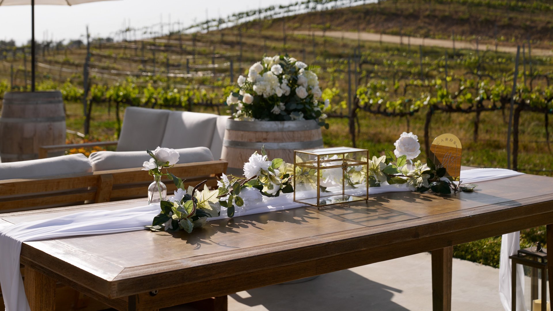 Rustic welcome and gift table at Danza del Sol Winery in Temecula, CA, decorated with greenery, candles, and vineyard views in the background.