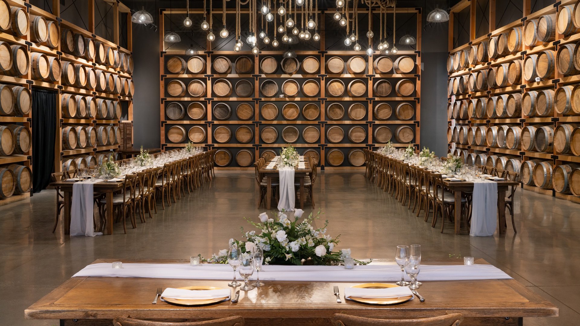 Center aisle view of the Barrel Room at Danza del Sol Winery, Temecula, CA, featuring wine barrels, rustic wood tables, and floral arrangements.