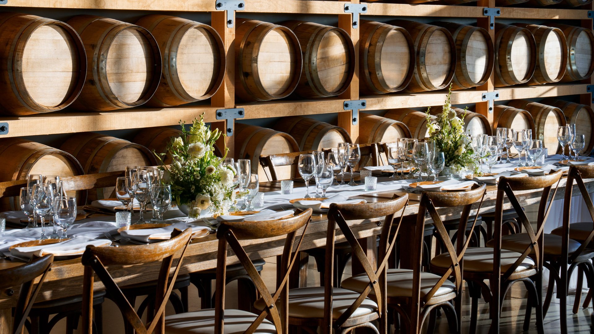 Reception table with floral centerpieces, glassware, and rustic wood chairs inside the Barrel Room at Danza del Sol Winery in Temecula, CA.