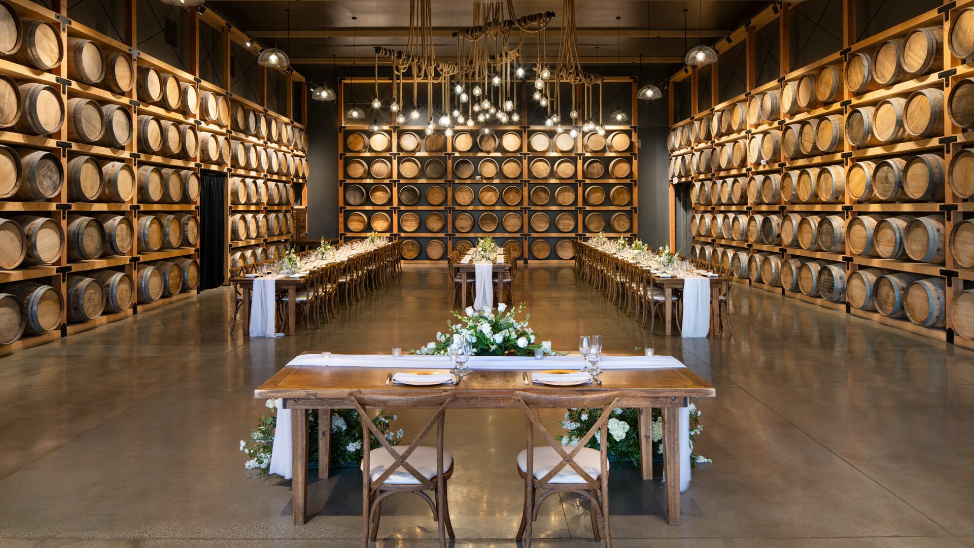 Rustic wedding reception inside Danza del Sol Winery Barrel Room with rows of wine barrels, romantic lighting, and farmhouse-style banquet tables.