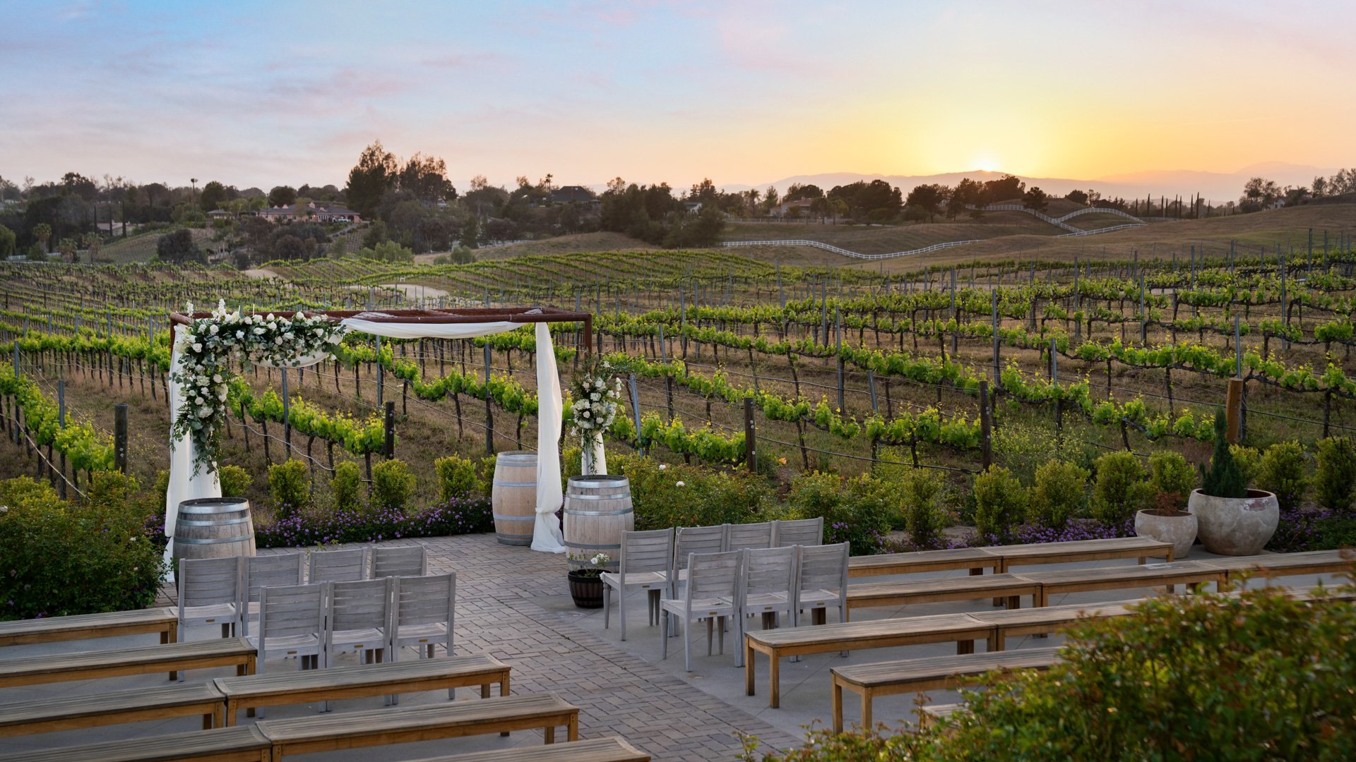 Outdoor vineyard wedding ceremony at Danza del Sol Winery in Temecula, CA, with floral arch, wine barrels, wooden benches, and sunset vineyard backdrop.