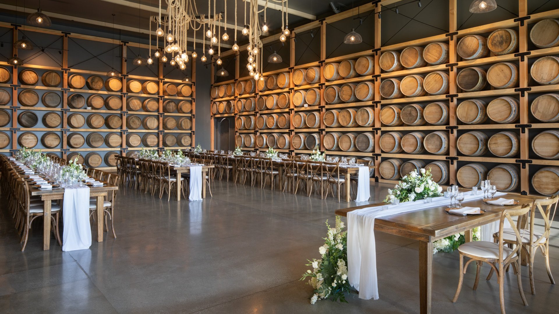 Sweetheart table with white floral arrangements inside the Barrel Room at Danza del Sol Winery, surrounded by wine barrels and banquet seating.