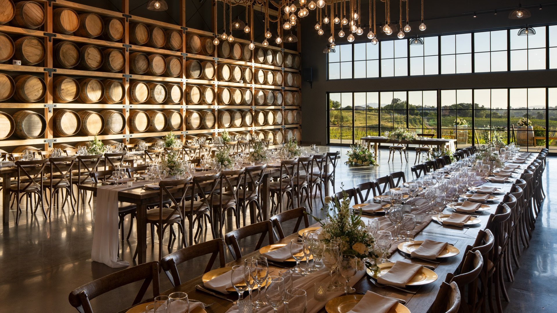 Winery wedding reception at Danza del Sol Winery in Temecula, CA, with rustic wood tables, wine barrel backdrop, and sunset light through glass doors.