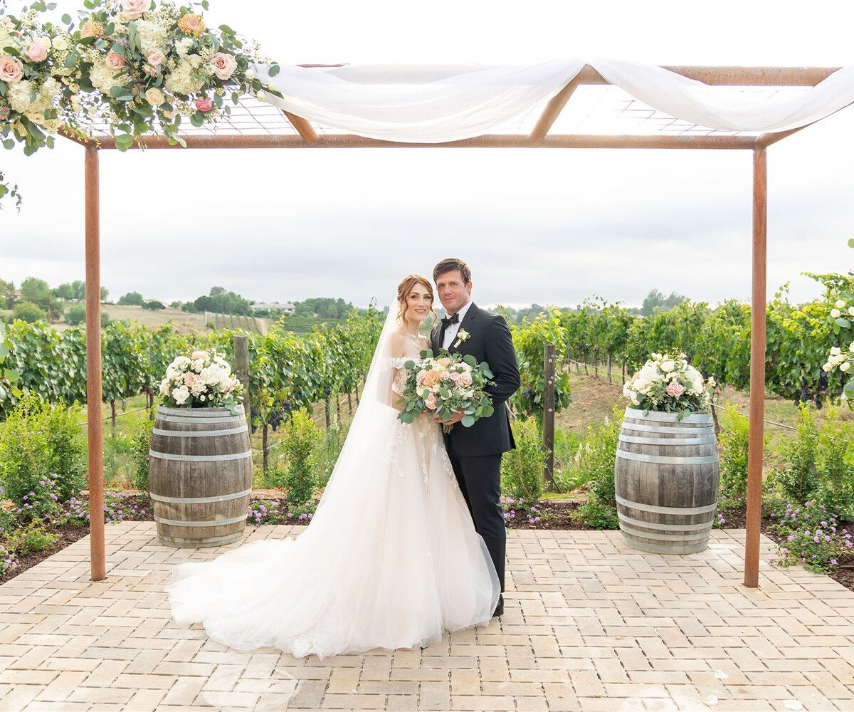Bride and groom portrait under pergola at Danza del Sol Winery Temecula with wine barrels, floral arrangements, and vineyard views