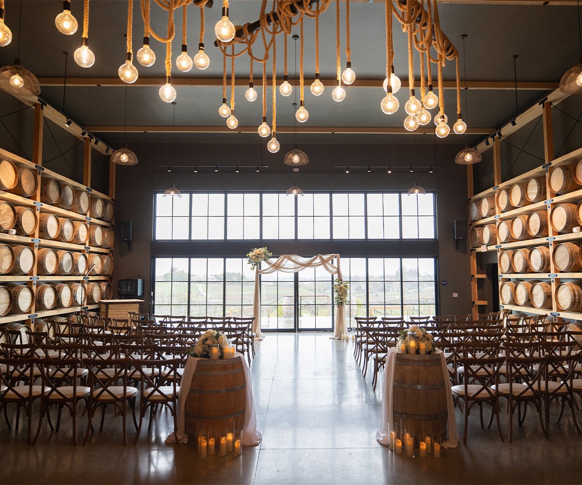 Indoor wedding ceremony at Danza del Sol Winery featuring barrel room with hanging Edison bulbs, wooden cross-back chairs, and wine barrel decor with candles lining the aisle towards floor-t