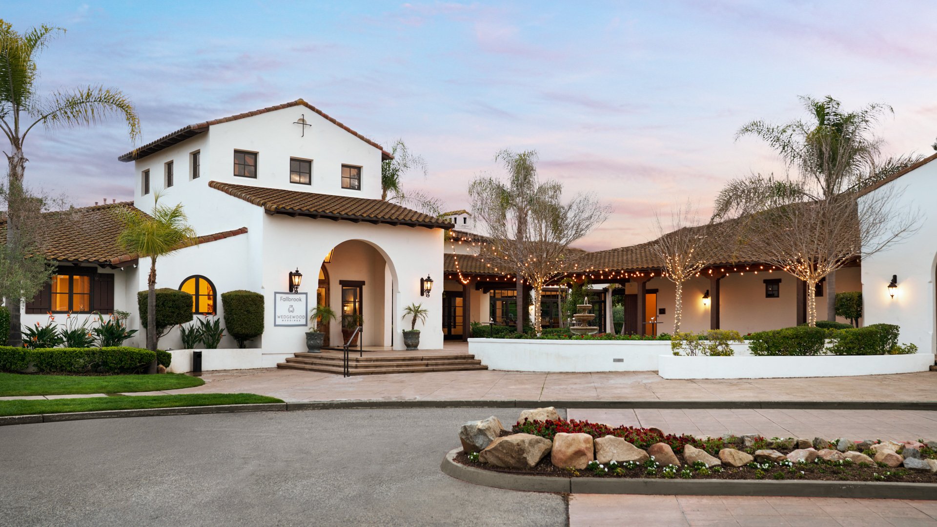 Spanish colonial style wedding venue exterior at Fallbrook Estate by Wedgewood Weddings featuring terracotta tile roof, white stucco walls, arched entrance and string lighting at dusk