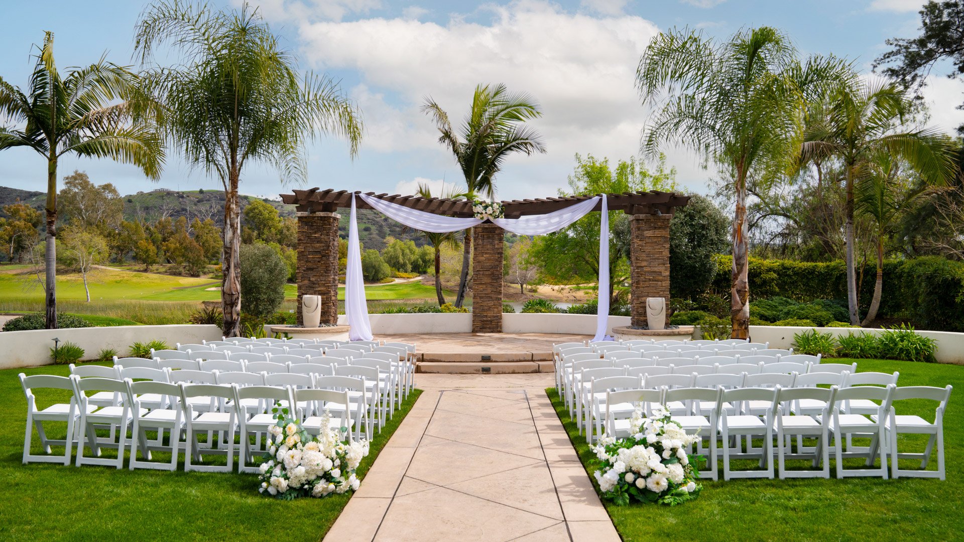 Elegant outdoor wedding ceremony setup at Fallbrook Estate by Wedgewood Weddings in Fallbrook, California featuring white chairs, stone archway with flowing fabric, palm trees and golf course views